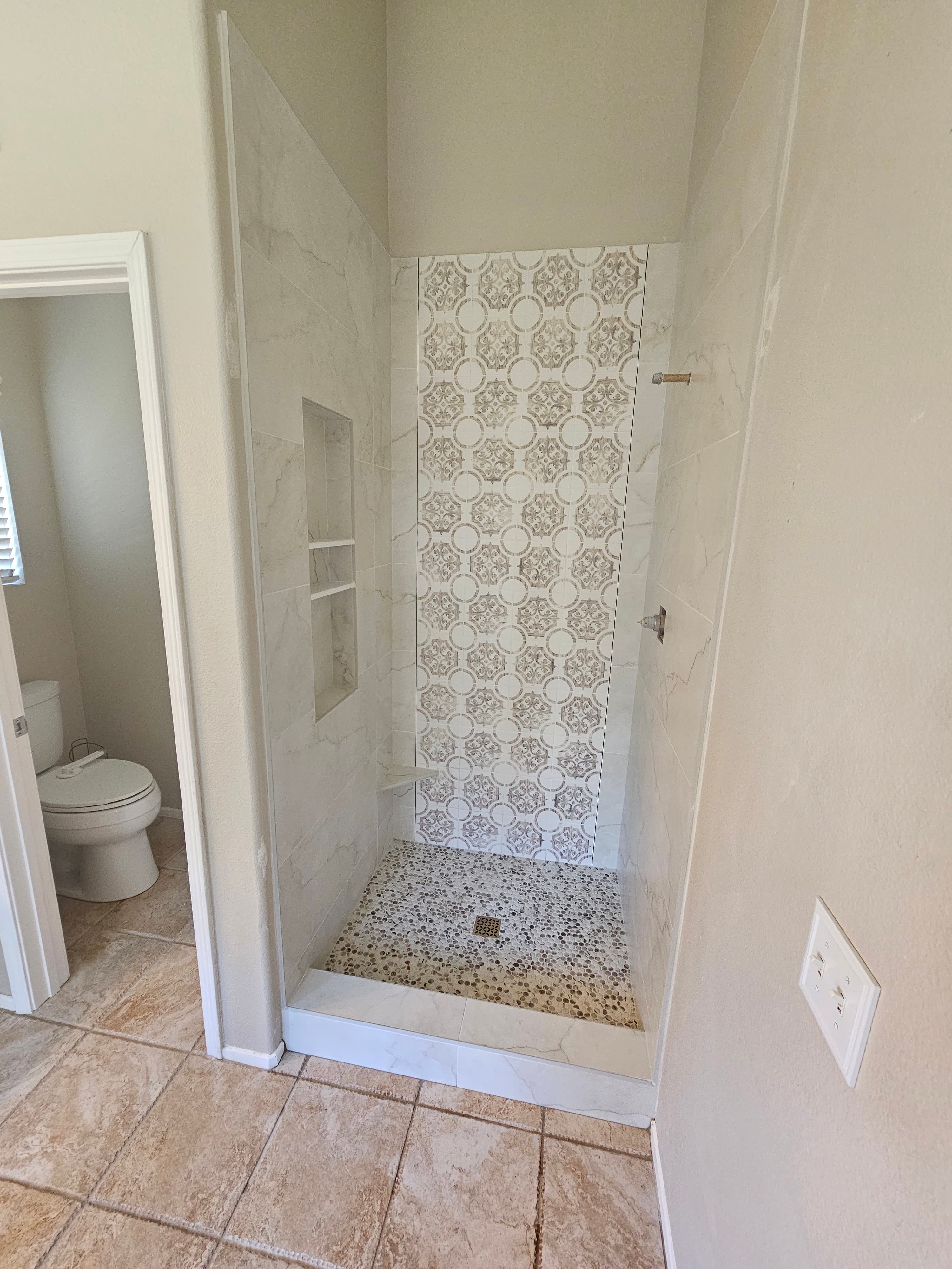 Walk-in Rancho Santa Margarita shower with marble side walls, geometric medallion-pattern back wall, and natural pebble mosaic floor